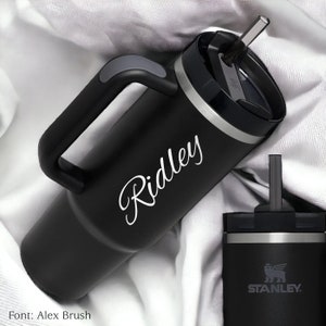 Pu&ograve; includere: Bicchiere termico nero con manico e cannuccia. Il nome "Ridley" &egrave; scritto in corsivo sul lato. &Egrave; visibile anche un bicchiere pi&ugrave; piccolo con il logo Stanley.