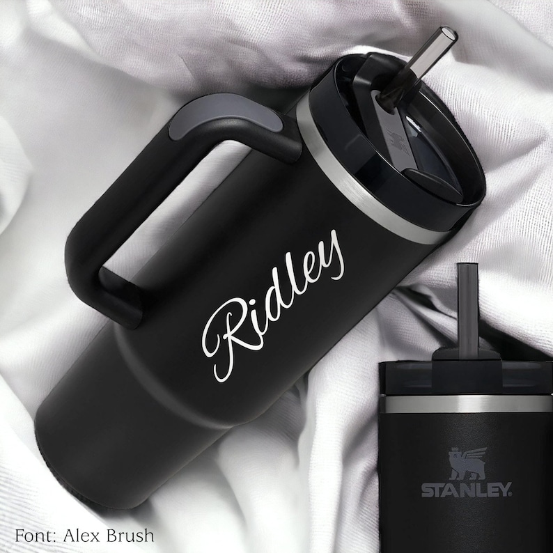 Pu&ograve; includere: Bicchiere termico nero con manico e cannuccia. Il nome "Ridley" &egrave; scritto in corsivo sul lato. &Egrave; visibile anche un bicchiere pi&ugrave; piccolo con il logo Stanley.