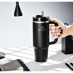 Pu&ograve; includere: Bicchiere Stanley nero con manico e cannuccia. Il bicchiere presenta il logo Stanley ed &egrave; tenuto da una persona. Il bicchiere &egrave; su una superficie bianca con una lampada nera e forniture per ufficio sullo sfondo.