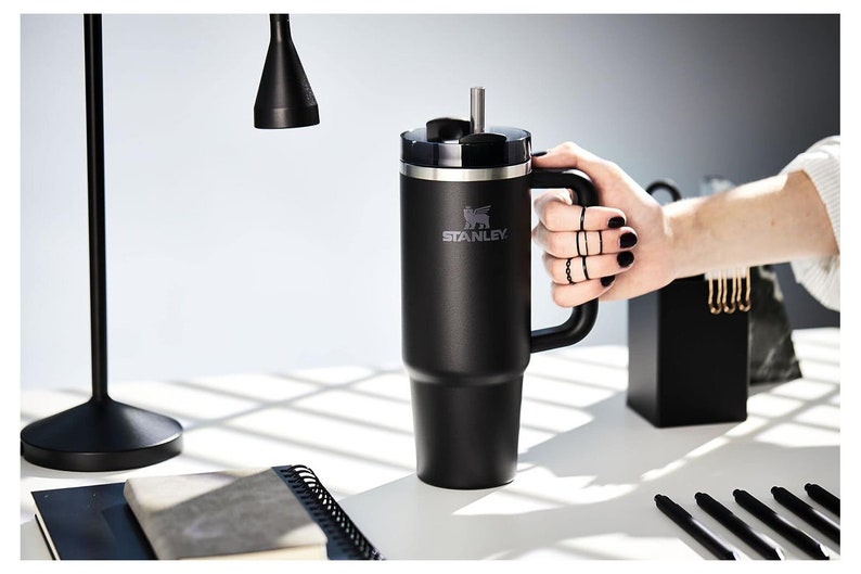 Pu&ograve; includere: Bicchiere Stanley nero con manico e cannuccia. Il bicchiere presenta il logo Stanley ed &egrave; tenuto da una persona. Il bicchiere &egrave; su una superficie bianca con una lampada nera e forniture per ufficio sullo sfondo.