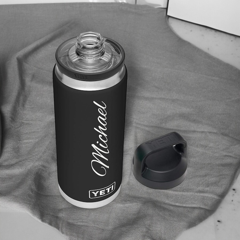 Puede incluir: Un enfriador de latas YETI negro con una tapa transparente y una tapa negra separada. El nombre "Michael" est&aacute; escrito en blanco en el lateral. El enfriador tiene un borde plateado y una base blanca. El logotipo de YETI es visible.