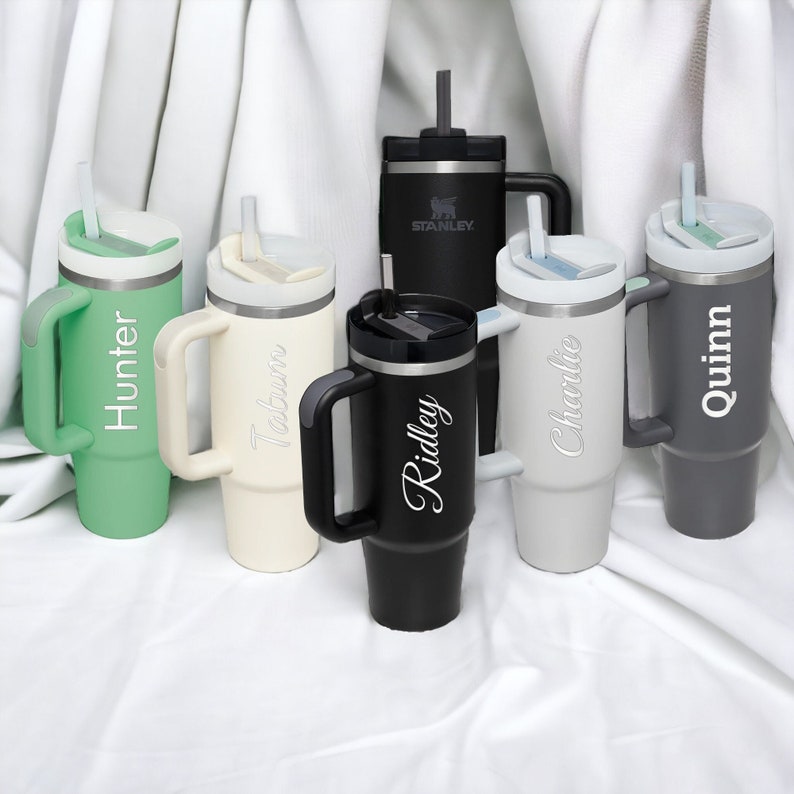 Pu&ograve; includere: Una collezione di bicchieri isolanti in vari colori, ciascuno con manico, coperchio e cannuccia. I bicchieri sono personalizzati con nomi come "Hunter", "Tatum", "Ridley", "Charlie" e "Quinn". Un bicchiere nero ha il logo "Stanley".