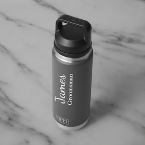 Puede incluir: Una botella de agua aislada gris con asa y tapa negras. La botella tiene el nombre "James" y la palabra "Groomsman" impresos en escritura blanca. La marca "YETI" est&aacute; impresa en la parte inferior.