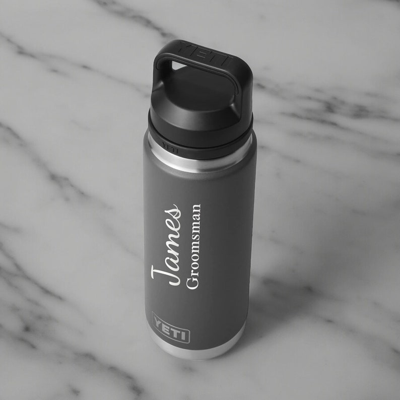 Puede incluir: Una botella de agua aislada gris con asa y tapa negras. La botella tiene el nombre "James" y la palabra "Groomsman" impresos en escritura blanca. La marca "YETI" est&aacute; impresa en la parte inferior.