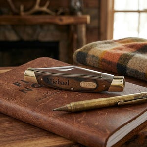May include: A personalized pocket knife with the name "Michael" engraved on the wooden handle, along with the "BUCK" brand logo. The knife rests on a leather-bound journal next to a brass pen. A plaid blanket and a fireplace are in the background.