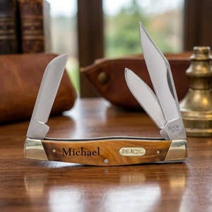 May include: A personalized pocket knife with a wooden handle and three stainless steel blades. The name "Michael" is engraved on the handle, along with the "BUCK 371+" logo. The knife is displayed open on a wooden surface.