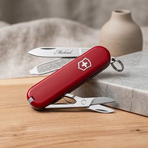May include: A red multi-tool pocket knife featuring a silver blade engraved with the name Michael, a nail file, and small scissors. The handle displays a white Swiss cross emblem, and a metal key ring is attached to one end of the tool.