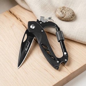 May include: A black folding pocket knife with a carabiner clip integrated into the handle. The blade features the name Michael engraved in a script font. The handle has a cutout design and a textured locking mechanism on the carabiner gate.