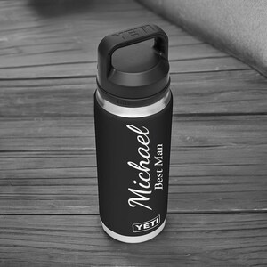 Puede incluir: Una botella de agua aislada negra y plateada con un asa negra. La botella tiene el nombre "Michael" y el texto "Best Man" en escritura blanca. La marca "YETI" es visible.