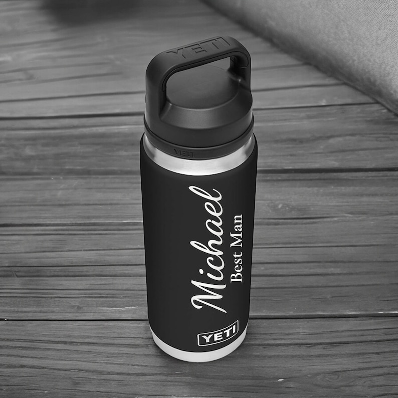 Puede incluir: Una botella de agua aislada negra y plateada con un asa negra. La botella tiene el nombre "Michael" y el texto "Best Man" en escritura blanca. La marca "YETI" es visible.