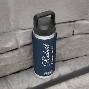 Puede incluir: Una botella de agua azul marino con asa y tapa negras. La botella tiene el nombre "Robert" en escritura blanca, con "Groomsman" debajo. La marca "YETI" est&aacute; impresa en la parte inferior.