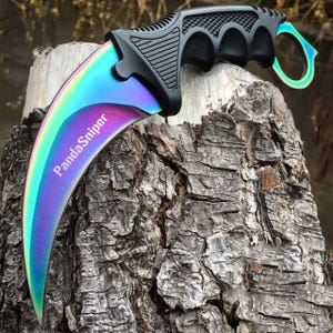 May include: A karambit knife with a rainbow-colored blade and a black handle. The blade curves inward, and the handle has finger grooves for grip. The text "PandaSniper" is printed on the blade. The knife rests on a textured, brown tree stump.