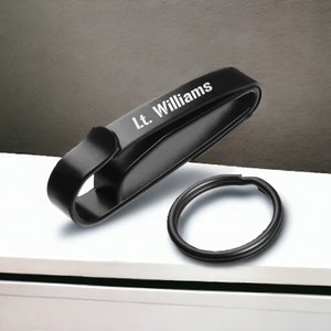 Op de afbeelding: Zwarte metalen sleutelhanger met een ring en een clip. De clip heeft de naam "Lt. Williams" gegraveerd.
