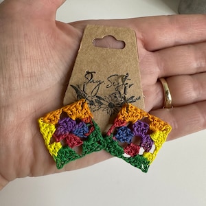 May include: A pair of crocheted earrings with a rainbow color pattern. The earrings are square shaped and have a small flower design in the center. The earrings are hanging from silver hooks.