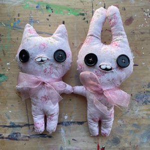 Op de afbeelding: Twee handgemaakte stoffen poppen. Eén is katvormig, de andere is konijnvormig. Beide poppen zijn gemaakt van roze bloemenstof, hebben grote zwarte knoopogen en kleine tanden. Elke pop heeft een roze strik en houdt elkaars hand vast.