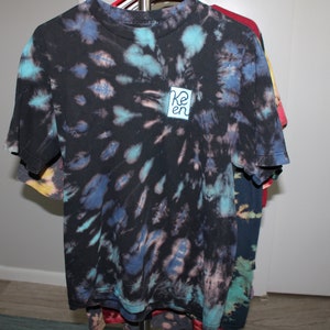 May include: A black tie-dye t-shirt with a blue and turquoise swirl pattern. The shirt has a small blue square patch with the word "Ken" written in white.
