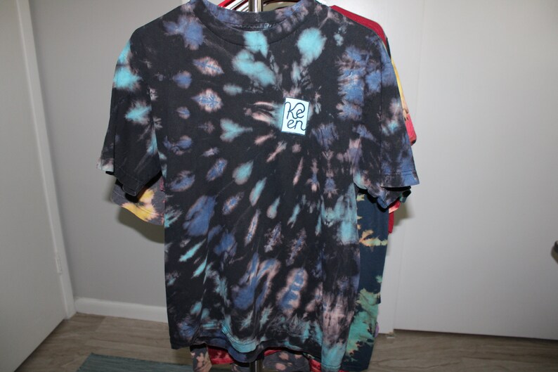 May include: A black tie-dye t-shirt with a blue and turquoise swirl pattern. The shirt has a small blue square patch with the word "Ken" written in white.