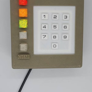 Può includere: Una tastiera grigia con un display bianco che mostra i numeri da 1 a 9 e 0. La tastiera ha un pulsante di cancellazione e tre luci colorate: rosso, arancione e verde.