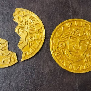 Puede incluir: Dos monedas de color dorado con diseños intrincados, una está rota por la mitad. Las monedas presentan una cara estilizada con patrones geométricos.