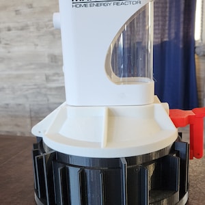 May include: A white 3D printed model of a Mr. Fusion home energy reactor from the film Back to the Future. The reactor has a clear plastic tube and a black base with a gear-like design.