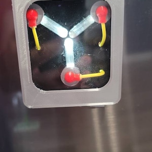 May include: A gray plastic frame with a black background and three red and yellow lights. The lights are arranged in a triangular pattern and are connected by white lines. The frame is a replica of the flux capacitor from the movie Back to the Future.