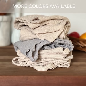 May include: A stack of folded muslin cloths featuring a crinkled, textured fabric in shades of beige and light gray. The top of the image includes the text MORE COLORS AVAILABLE in white capital letters against a semi-transparent gray bar.