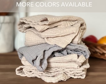 May include: A stack of folded muslin cloths featuring a crinkled, textured fabric in shades of beige and light gray. The top of the image includes the text MORE COLORS AVAILABLE. The cloths are layered on top of each other in a neat pile.