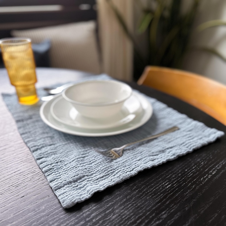 May include: A light blue rectangular placemat with a crinkled, textured surface. On the placemat sits a white bowl stacked on a white plate, a silver fork, and a tall, ribbed amber-colored glass. The items are arranged on a dark wooden table.