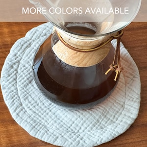 May include: A glass pour-over coffee maker with a light wood collar and a brown leather tie, filled with dark coffee, resting on a round, textured, light gray fabric mat. The text MORE COLORS AVAILABLE is visible at the top of the image.