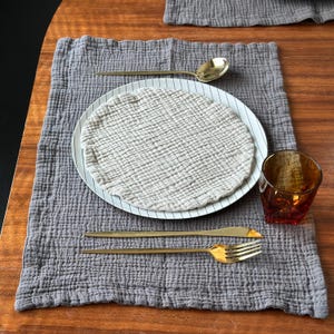 May include: A grey textured fabric placemat featuring a white plate with thin black stripes, topped with a round beige muslin cloth. The setting includes a gold spoon, a gold knife, a gold fork, and an amber-colored glass tumbler.
