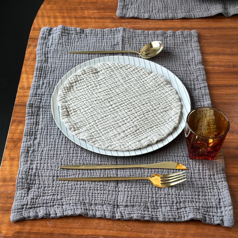 May include: A grey textured fabric placemat featuring a white plate with thin black stripes, topped with a round beige muslin cloth. The setting includes a gold spoon, a gold knife, a gold fork, and an amber-colored glass tumbler.