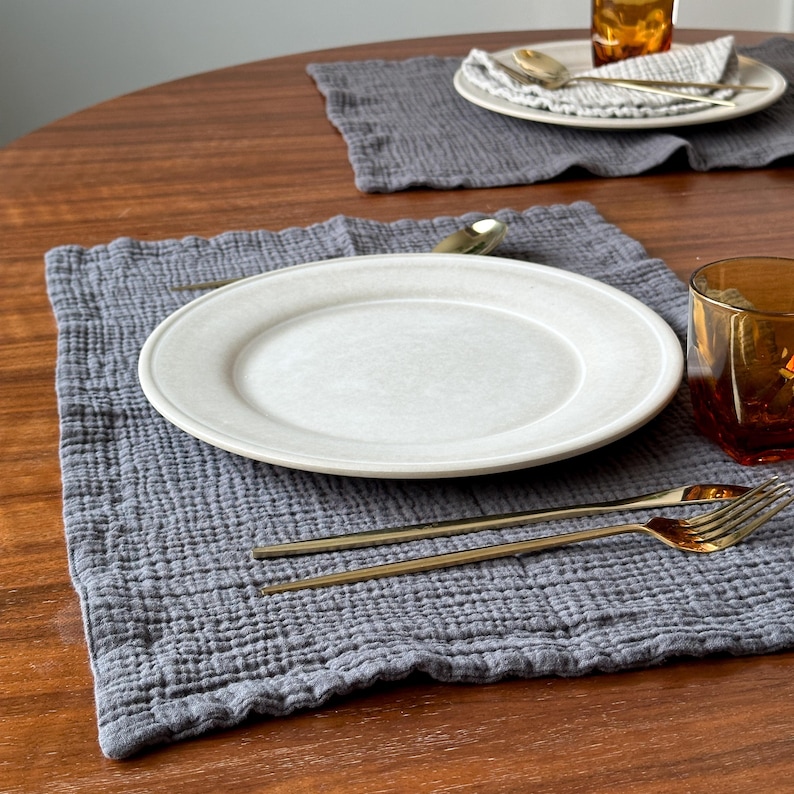 May include: A square, dark gray, textured cotton muslin placemat featuring a smooth, off-white ceramic dinner plate centered on top. A gold-toned fork and knife are placed on the fabric, alongside a clear amber glass tumbler.