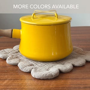 May include: A bright yellow enamel saucepan with a matching lid and a light brown wooden handle. The pot sits on a beige, scalloped-edge fabric trivet. The text "MORE COLORS AVAILABLE" is printed in white at the top of the image.