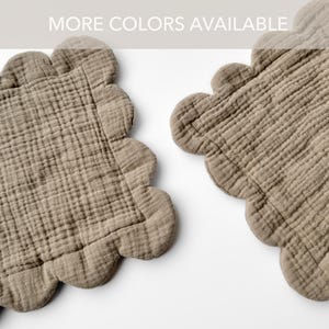 May include: Two beige, scalloped-edge placemats made of a textured fabric. The placemats are square with a wavy border. The text "MORE COLORS AVAILABLE" is at the top of the image.