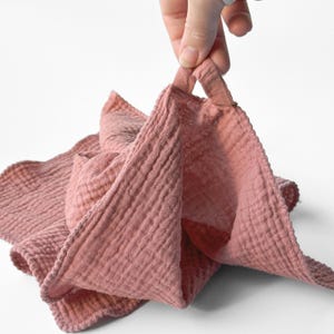 May include: A dusty rose-colored, textured washcloth with a loop for hanging. The cloth is made of a soft, crinkled fabric, and is held up by a hand against a white background. The washcloth is folded and appears to be new.