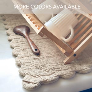 May include: A beige scalloped-edge drying mat with a wooden dish rack, a dark brown wooden spoon, and a white ribbed mug. The text MORE COLORS AVAILABLE is visible at the top. The mat has a textured, layered fabric surface and a soft, wavy border.