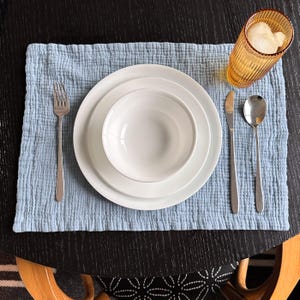 May include: A light blue textured cotton placemat featuring a white ceramic bowl stacked on a white dinner plate. A silver fork is placed on the left, while a silver knife and spoon sit on the right. An amber ribbed glass with ice is included.