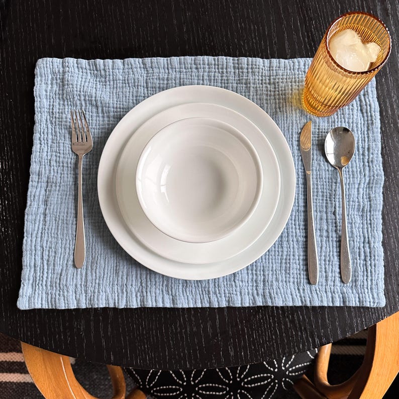 May include: A light blue textured cotton placemat featuring a white ceramic bowl stacked on a white dinner plate. A silver fork is placed on the left, while a silver knife and spoon sit on the right. An amber ribbed glass with ice is included.
