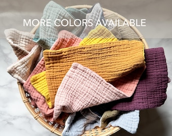 May include: A collection of folded muslin cloths in various colors including mustard yellow, dusty pink, deep purple, light blue, and beige, arranged inside a round woven wicker basket. The text MORE COLORS AVAILABLE is printed on the image.