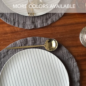 May include: White plates with vertical black stripes are set on gray placemats. A gold spoon and fork are placed on the plates. The text "MORE COLORS AVAILABLE" is at the top. The table is made of wood.