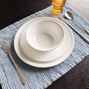 May include: A white ceramic dinnerware set consisting of a bowl, a small plate, and a large plate stacked together. The set is accompanied by a silver fork, a silver knife, a silver spoon, and an amber-colored ribbed glass on a light blue placemat.
