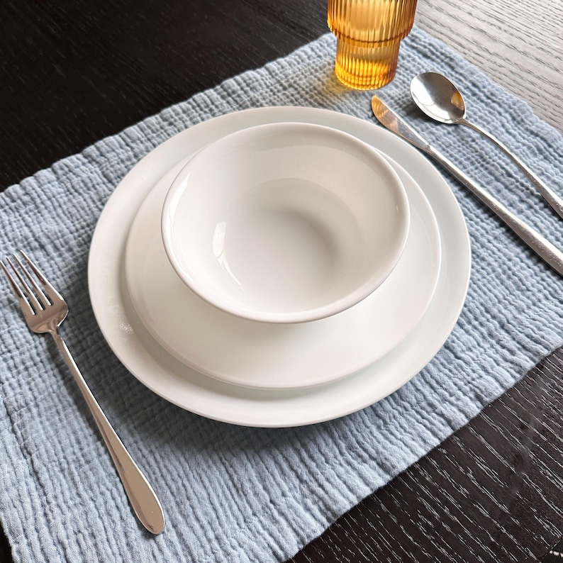 May include: A white ceramic dinnerware set consisting of a bowl, a small plate, and a large plate stacked together. The set is accompanied by a silver fork, a silver knife, a silver spoon, and an amber-colored ribbed glass on a light blue placemat.