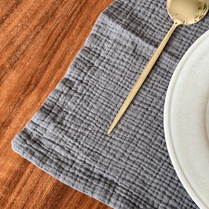 May include: A dark gray rectangular placemat with a textured, crinkled fabric surface. A polished gold-toned metal spoon with a long, slender handle rests diagonally on the fabric. A portion of a white ceramic plate is visible on the right side.