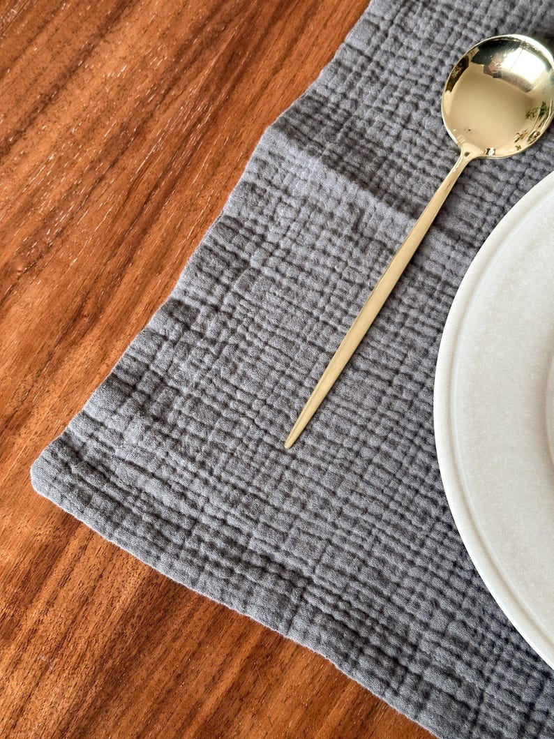 May include: A dark gray rectangular placemat with a textured, crinkled fabric surface. A polished gold-toned metal spoon with a long, slender handle rests diagonally on the fabric. A portion of a white ceramic plate is visible on the right side.