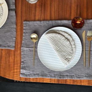 May include: A grey textured fabric placemat featuring a white plate with thin black horizontal stripes. A folded beige muslin cloth rests on the plate. A gold spoon, a gold knife, a gold fork, and a small amber glass are arranged on the placemat.