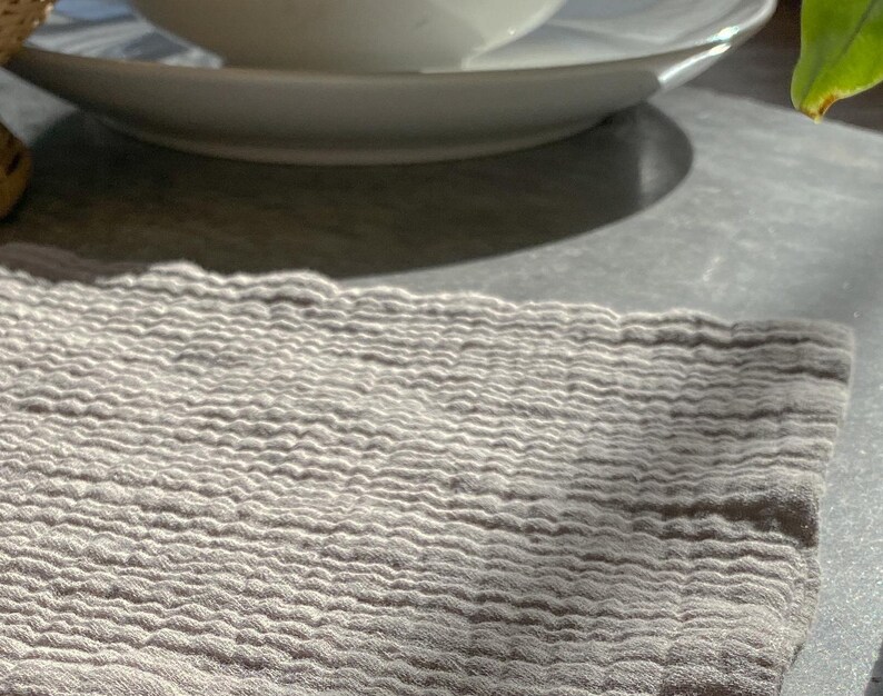 May include: A light gray, rectangular textile cloth featuring a distinct, multi-layered crinkled texture. The fabric displays a consistent, fine-grained, puckered surface pattern throughout, creating a soft, dimensional appearance across the entire piece.