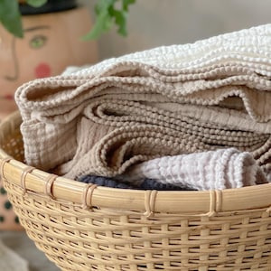May include: A woven wicker basket filled with folded, textured linen towels in various neutral colors, including white, beige, and taupe. The towels have a soft, crinkled appearance. A decorative pot with a face design is in the background.