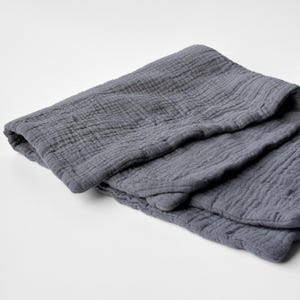 May include: A folded, slate-gray muslin blanket. The blanket has a crinkled texture and is folded in thirds, with the edges neatly tucked. The fabric appears soft and lightweight, suitable for a baby or as a decorative throw.