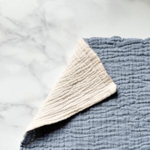 May include: A double-layered muslin cloth featuring a textured, crinkled surface. The fabric is colored in a muted dusty blue on the top side, while the folded corner reveals a contrasting, solid off-white or cream-colored underside.