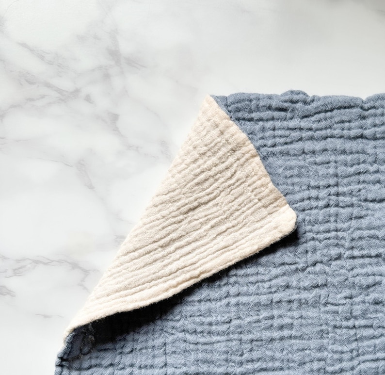 May include: A double-layered muslin cloth featuring a textured, crinkled surface. The fabric is colored in a muted dusty blue on the top side, while the folded corner reveals a contrasting, solid off-white or cream-colored underside.
